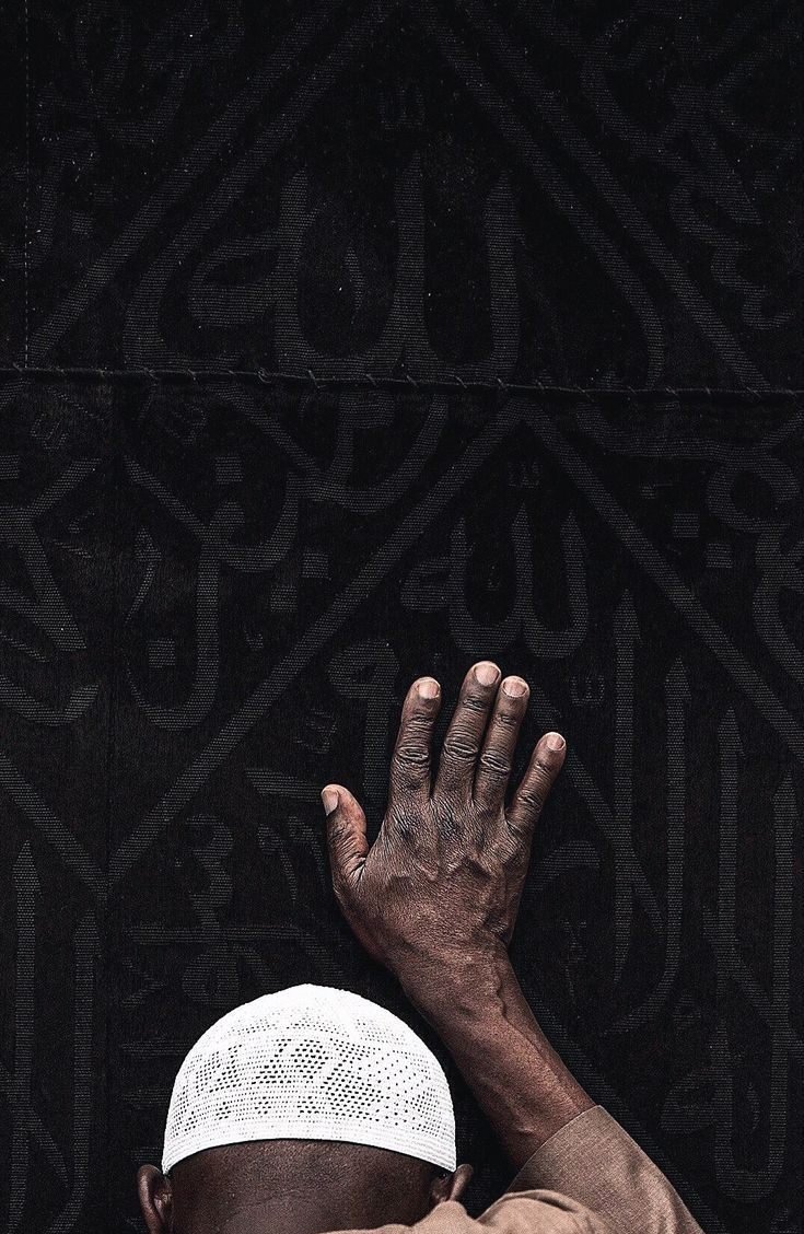 A worshipper resting a hand against the embroidered black kiswah of the Kaaba