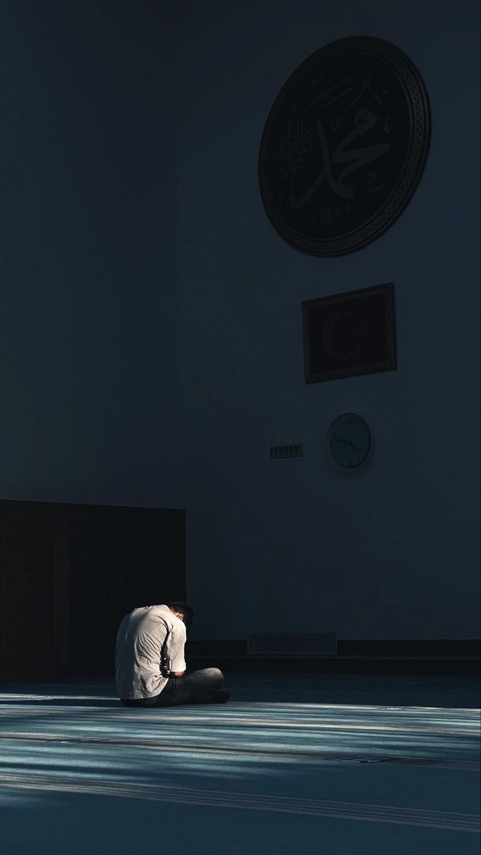 A person sitting alone in quiet prayer on a mosque carpet, lit by shafts of daylight